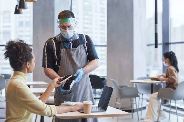 Cintura para arriba retrato de camarero con protector facial y guantes mientras toma el pago en la cafetería con medidas de seguridad de covid, espacio de copia