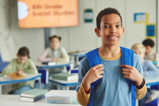 Cintura para arriba retrato de colegial joven negro con mochila mirando a la cámara mientras está de pie en classro