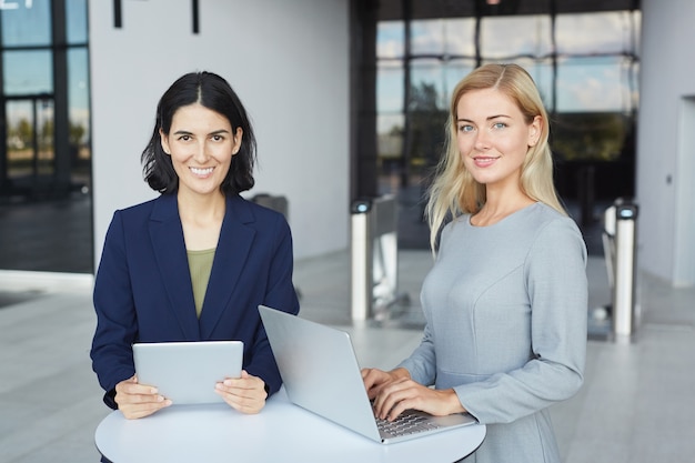 Cintura para arriba retrato de dos mujeres empresarias exitosas sonriendo a la cámara mientras está de pie en un escritorio en el edificio de oficinas y sosteniendo el portátil,