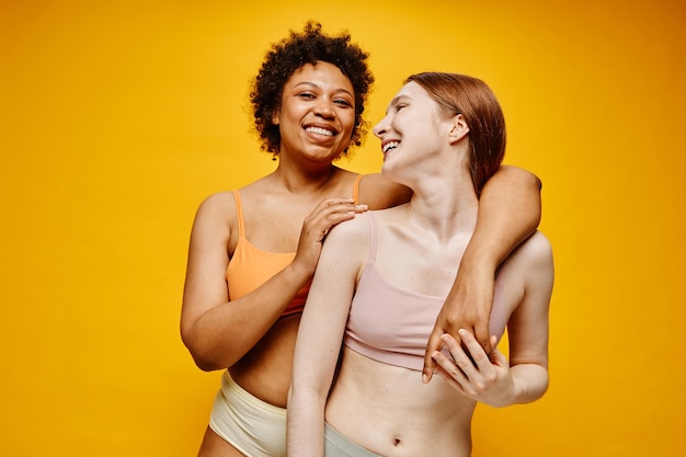 Cintura para arriba retrato de dos mujeres jóvenes despreocupadas de piel oscura y piel clara abrazándose mientras están de pie de nuevo