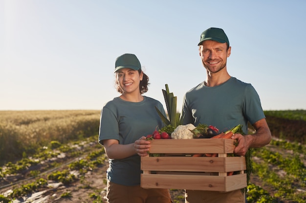 Cintura para arriba retrato de dos trabajadores sosteniendo una caja de verduras y sonriendo a la cámara mientras está de pie en una plantación de hormigas al aire libre, espacio de copia