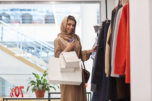 Cintura para arriba retrato de elegante mujer de Oriente Medio mientras está de pie junto a los percheros y disfruta de las compras en el centro comercial, espacio de copia