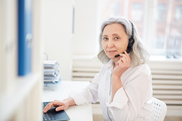 Cintura para arriba retrato de empresaria senior hablando al micrófono y mirando mientras trabaja con la computadora portátil en el interior de la oficina