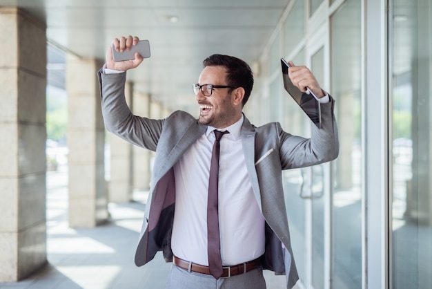 Cintura para arriba retrato del empresario celebrando el éxito. en una tableta de mano y en otro teléfono inteligente. centro de negocios exterior.