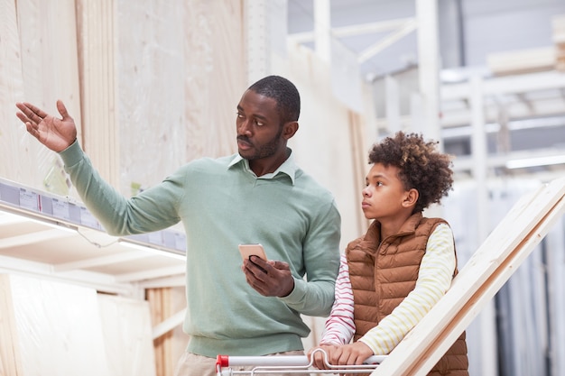 Cintura para arriba retrato de hombre afroamericano apuntando a tablas de madera mientras compra con el hijo en la ferretería, espacio de copia