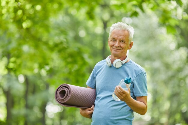 Cintura para arriba retrato de hombre senior activo sosteniendo una estera de yoga y una botella de agua mirando a la cámara