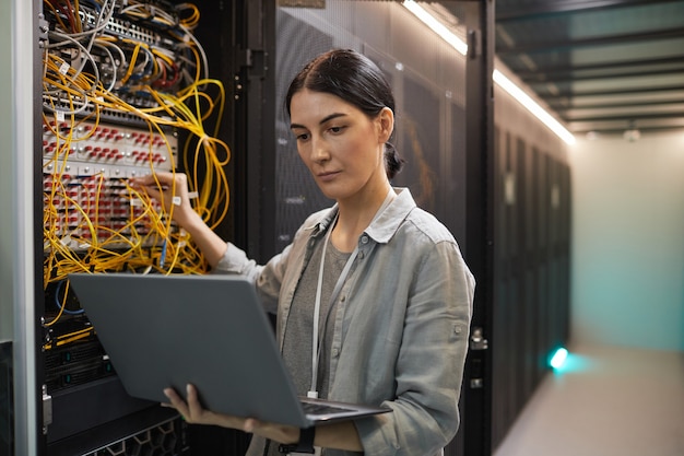 Cintura para arriba retrato de ingeniero de redes femenino conectando cables en el gabinete del servidor mientras trabaja con la supercomputadora en el centro de datos, espacio de copia