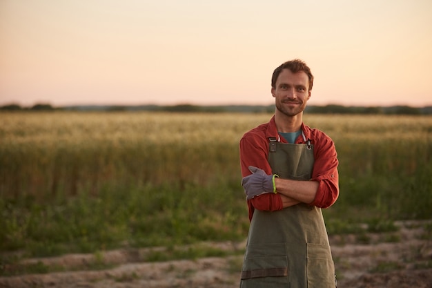 Cintura para arriba retrato de joven agricultor posando con confianza con los brazos cruzados mientras está de pie en el campo al atardecer y sonriendo a la cámara, espacio de copia