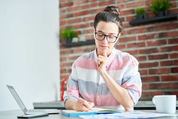 Cintura para arriba retrato de joven moderna estudiando en casa y escribiendo en el cuaderno