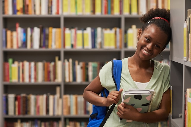 Cintura para arriba retrato de joven mujer afroamericana de pie en la biblioteca de la escuela y sonriendo a la cámara ...