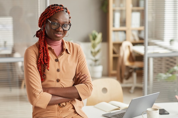 Cintura para arriba retrato de joven mujer afroamericana sonriendo a la cámara mientras se inclina sobre el escritorio en el interior de la oficina moderna, espacio de copia