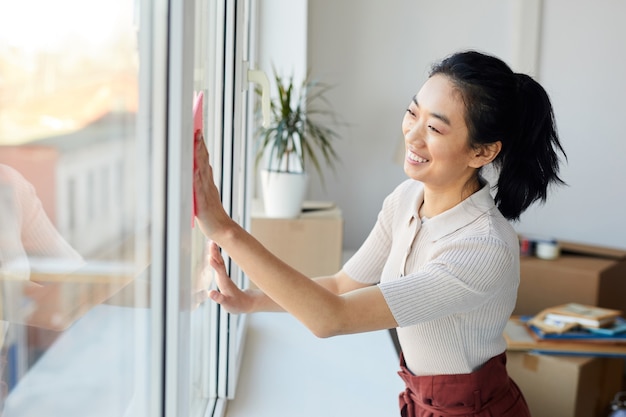 Cintura para arriba retrato de joven mujer asiática lavando ventanas mientras disfruta de la limpieza de primavera en casa o apartamento