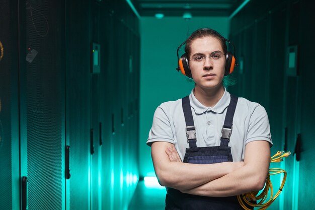 Foto cintura para arriba retrato de joven técnico de red mirando a la cámara mientras está de pie con los brazos cruzados en la sala de servidores, espacio de copia