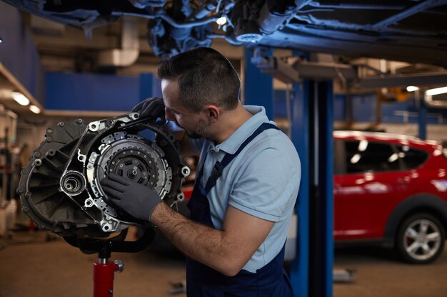 Foto cintura para arriba retrato de mecánico de automóviles muscular inspeccionando la caja de cambios en el taller de reparación de automóviles, espacio de copia