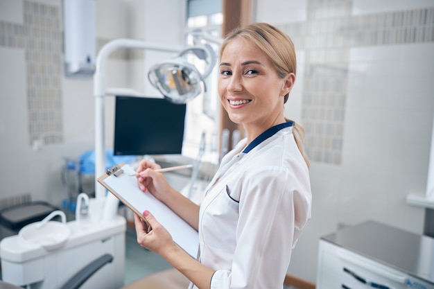 Cintura para arriba retrato de mujer bonita alegre escribiendo en el portapapeles mientras está de pie cerca de la silla dental