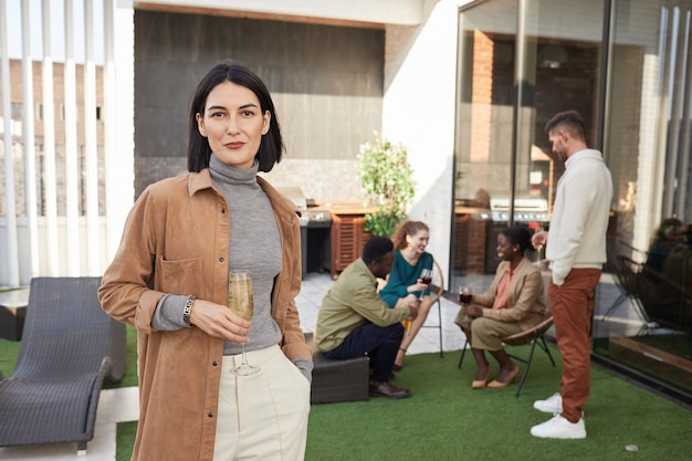 Cintura para arriba retrato de mujer joven contemporánea y sonriendo mientras está de pie en la terraza durante la fiesta al aire libre,