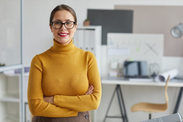 Cintura para arriba retrato de mujer sonriente arquitecto de pie con los brazos cruzados y mientras posa en el lugar de trabajo en la oficina,