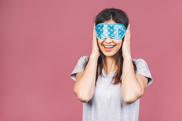 Cintura para arriba retrato de niña alegre sonriente en máscara para dormir. Atractiva mujer divertida en elegante pijama de pie y mirando a otro lado. Aislado sobre fondo rosa