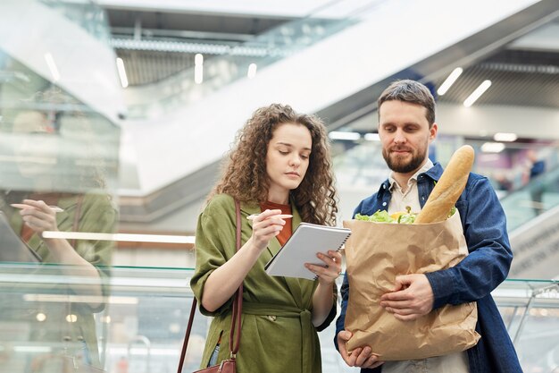 Cintura para arriba retrato de pareja moderna comprobación de lista de compras mientras compra alimentos en el supermercado