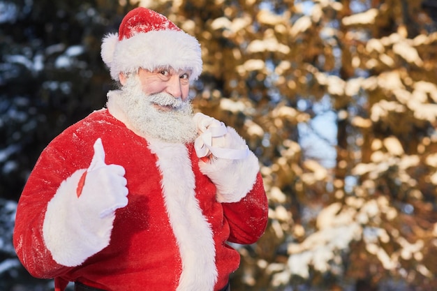 Cintura para arriba retrato de santa claus tradicional llevando saco con regalos al aire libre y mostrando los pulgares