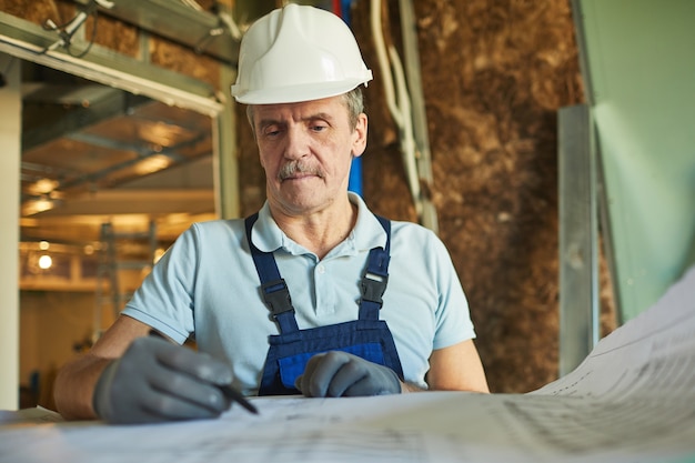 Cintura para arriba retrato de trabajador de la construcción senior con casco mientras mira los planos de planta mientras se renueva la casa, espacio de copia