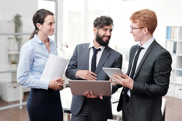 Cintura para arriba retrato de tres jóvenes empresarios discutiendo el trabajo y sonriendo mientras está de pie en la oficina moderna, espacio de copia