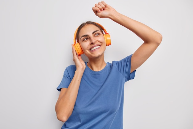 Foto cintura para arriba tiro de alegre mujer soñadora baila con ritmo de música usa auriculares estéreo naranjas en las orejas vestidas con una camiseta azul casual aislada sobre fondo blanco concepto de personas y pasatiempos