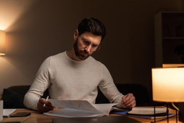 Cintura arriba vista de retrato del hombre barbudo con suéter blanco leyendo y escribiendo cartas en casa durante la noche acogedora. Foto de stock