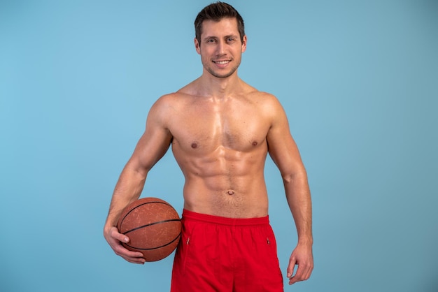 Cintura para arriba vista de retrato del joven posando con pelota sobre fondo azul aislado mientras juega baloncesto