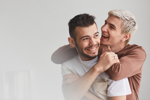 Cintura franca para arriba retrato de una pareja gay despreocupada abrazándose en el interior y riendo felizmente mientras posa contra el espacio blanco, copia