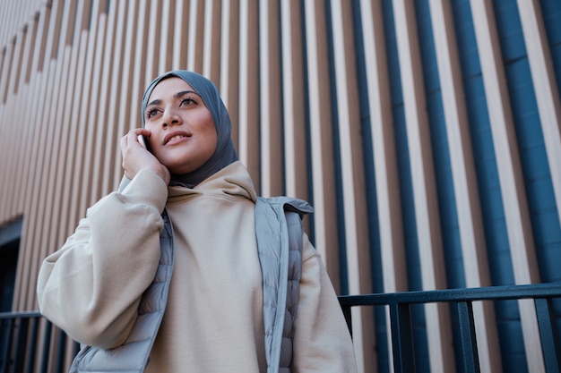 Cintura gráfica para arriba retrato de mujer moderna del Medio Oriente hablando por teléfono inteligente en la ciudad, espacio de copia