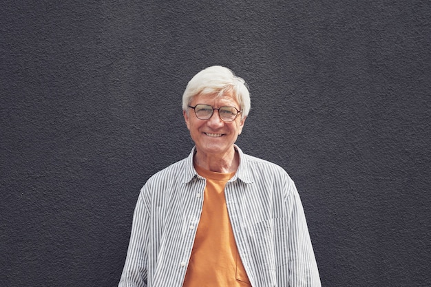 Cintura mínima para arriba retrato de hombre senior moderno mirando a cámara y sonriendo mientras está de pie junto a la pared negra al aire libre, espacio de copia