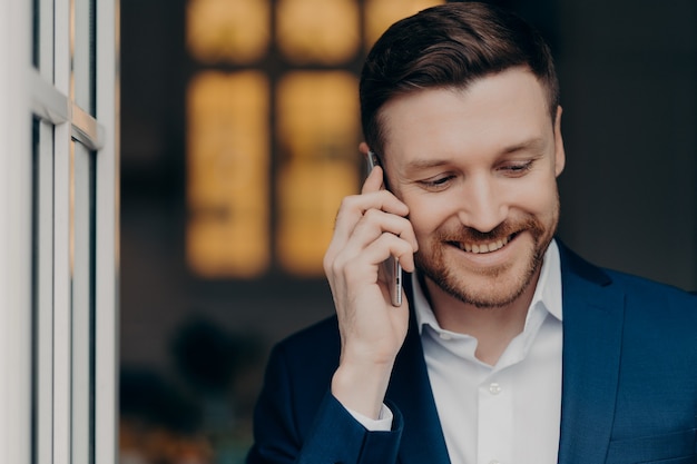 Cintura para cima de sorridente empresário barbudo de terno tendo conversa móvel corporativa, desfrutando de uma boa conversa enquanto está dentro de casa e olhando para baixo com expressão feliz. Conceito de negócios