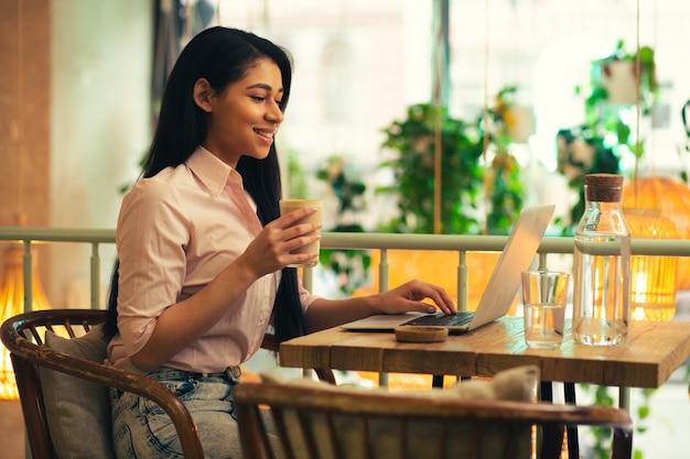 Cintura para cima de uma estudante confiante de cabelos compridos segurando um copo de café com leite e sorrindo enquanto usa seu laptop no café