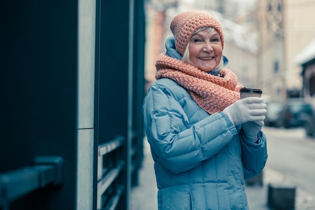 Cintura para cima de uma mulher idosa positiva em pé na rua com uma xícara de café e sorrindo. Roupas quentes de inverno na mulher