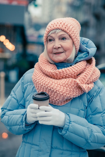 Cintura para cima de uma senhora idosa calma em roupas quentes de inverno em pé ao ar livre com uma xícara de café e olhando para longe