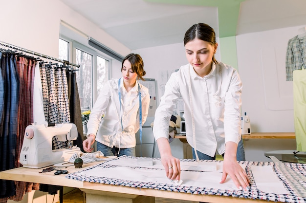 Cintura para cima o retrato de duas jovens mulheres bonitas alfaiates trabalhando com padrões de roupas em tecido enquanto costura calças clássicas no ateliê, cópia espaço