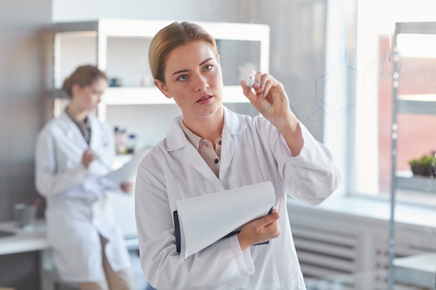 Cintura para cima retrato de jovem cientista escrevendo na parede de vidro e segurando a prancheta enquanto está no laboratório médico, copie o espaço