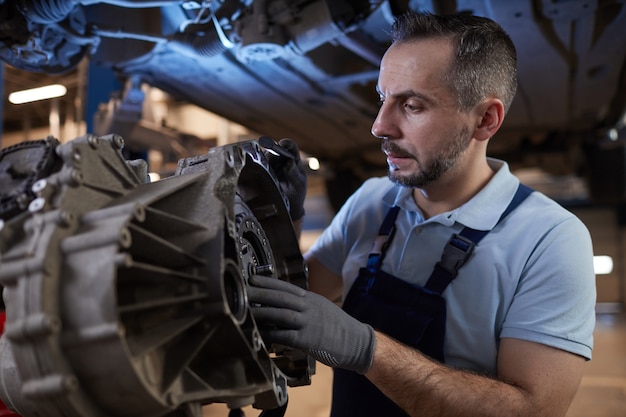 Foto cintura para cima retrato de mecânico de automóveis musculoso inspecionando peça de carro em oficina mecânica, copie o espaço