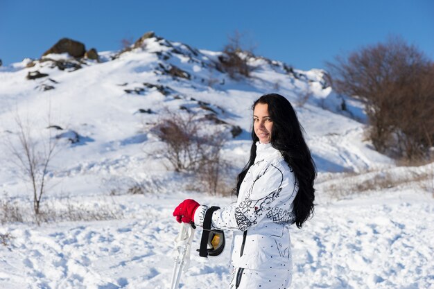 Cintura para cima retrato de mulher com longos cabelos escuros, vestindo roupa de esqui branca e sorrindo por cima do ombro para a câmera, tirando uma folga do esqui na montanha coberta de neve em um dia ensolarado com luz do sol