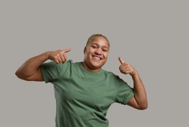 Cintura para cima retrato de mulher de raça mista andrógina sorrindo para a câmera enquanto posa em um fundo cinza no estúdio e mostrando os polegares para cima, copie o espaço