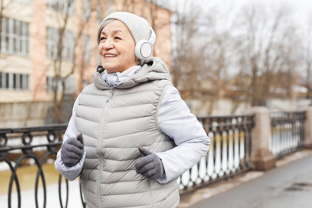 Cintura para cima, retrato de mulher sênior ativa correndo ao ar livre no inverno e sorrindo feliz, copie o espaço