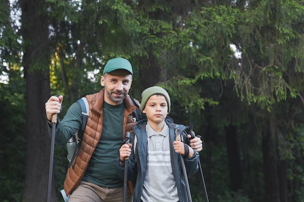 Cintura para cima retrato de pai e filho felizes caminhando juntos e enquanto caminhavam na floresta com bastões de caminhada, copie o espaço