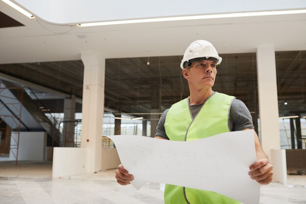 Cintura para cima retrato de trabalhador da construção civil profissional segurando plantas e olhando para longe enquanto está em um prédio de escritórios,