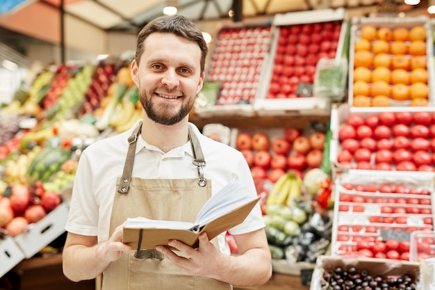 Cintura para cima, retrato de um homem barbudo usando avental e sorrindo enquanto está de pé ao lado de uma barraca de frutas e vegetais no mercado dos fazendeiros