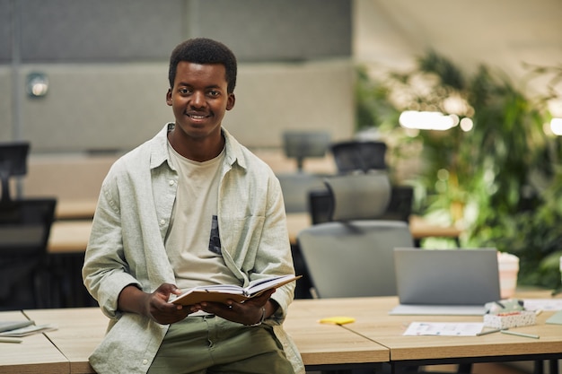 Cintura para cima, retrato de um jovem afro-americano sorrindo, segurando o planejador e apoiado na mesa em um escritório moderno, copie o espaço