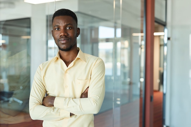 Cintura para cima, retrato de um jovem empresário afro-americano vestindo uma camisa amarela, posando com confiança e olhando para a câmera de pé com os braços cruzados no interior do escritório de vidro, copie o espaço