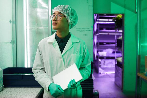 Cintura para cima, retrato de um jovem trabalhador asiático vestindo roupas protetoras em pé em um laboratório biológico iluminado por luz verde, copie o espaço