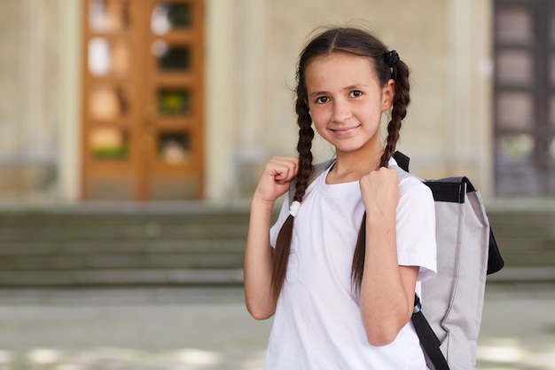 Cintura para cima retrato de uma colegial bonita carregando uma mochila e sorrindo para a câmera enquanto posa ao ar livre contra o prédio da escola, copie o espaço