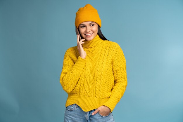 Cintura para cima, retrato de uma jovem com uma camisola de malha amarela falando no smartphone e desviando o olhar com um sorriso isolado no azul